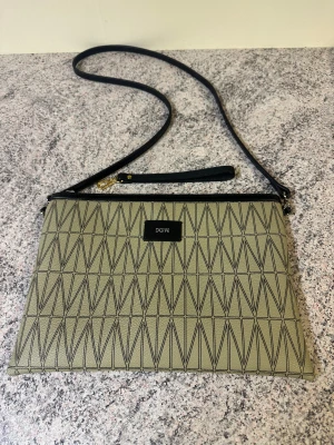 Beige clutch från Dagmar med mönster - Snygg beige clutch från Dagmar med geometriskt mönster i svart. Väskan har en smal svart axelrem, dragkedja upptill och flera fack inuti. Toppen skick invändigt och utvändigt. Perfekt för dig som gillar stilrena accessoarer med lite edge.