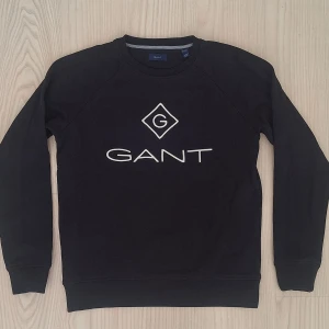 Svart sweatshirt från GANT 158/164 - Nyskick, använd ett fåtal gånger. Svart sweatshirt från GANT med vit logotyp framtill. Klassisk rund halsringning och långa ärmar. Mjuk och skön bomullsblandning, perfekt för en clean och stilren look.