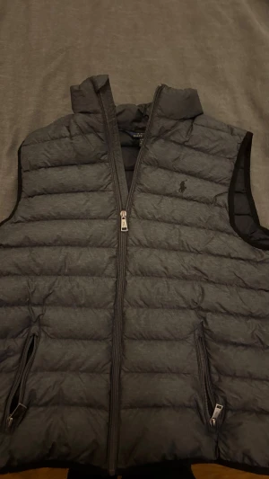 Grå dunväst från Polo Ralph Lauren - Snygg grå dunväst från Polo Ralph Lauren med horisontella quiltade paneler och diskret logga på bröstet. Västen har två praktiska fickor med dragkedja. Perfekt för lager-på-lager och enkel att matcha med din stil.size 14-16 motsvarar xs kanske s.skriv gärna om ni har grågor.priset kan diskuteras.