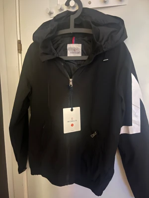 Moncler vindjacka  - Säljer en Moncler vindjacka i ett otrolig skick. Jackan är svart med en stilren vit loga på armen med bokstav M. 