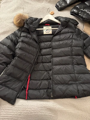 Svart dunjacka med fuskpäls från JOTT - Snygg svart dunjacka från JOTT med avtagbar huva och fuskpälsdetalj. Jackan har quiltad design, dragkedja framtill och röda detaljer på insidan. Perfekt för kalla dagar och har en klassisk badge på ärmen.