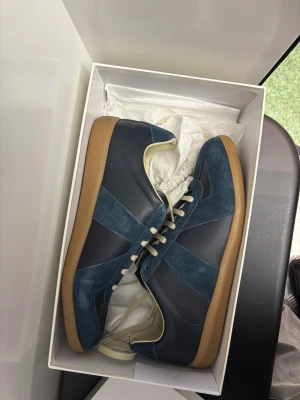 Maison Margiela blå sneakers - Maison Margiela sneakers i mörkblå mocka och skinn med beige detaljer och snörning. Snygg kontrast mellan material och färger, samt klassisk gummisula i brunt. Ikonisk logotyp på plösen och undersidan. Perfekt för dig som gillar stilrena och exklusiva sneakers.