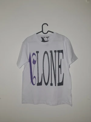 Palm Angels - Vit t-shirt med stor svart och lila VLONE-print på framsidan och en stor lila V med flames och svart Palm Angels-tryck på ryggen. Klassisk rund hals och korta ärmar. Streetwear-vibe och riktigt snygg statement piece.
