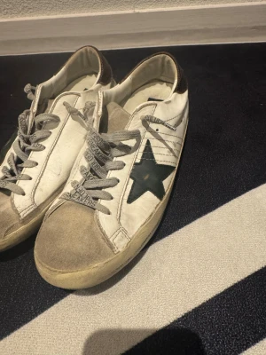 Golden Goose Superstar sneakers - Golden Goose Superstar sneakers i vitt skinn med grå mockadetaljer på tån och grön stjärna på sidan. Skorna har grå snörning med tryckt text och bruna detaljer vid hälen. Platt sula och rund tå ger en klassisk streetstil.