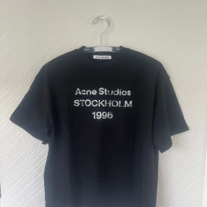 Svart Acne Studios t-shirt S Stockholm - Svart t-shirt från Acne Studios med vit text 'Acne Studios STOCKHOLM 1996' på bröstet. Klassisk rund hals och korta ärmar. Materialet är mjuk bomull och passformen är normal. Perfekt för dig som gillar stilrena och ikoniska plagg med streetkänsla.