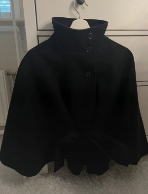 Svart cape-kappa med bälte - Svart cape-kappa från Gina tricot  med hög krage och tre stora knappar framtill. Kappan har ett brett bälte som knyts i midjan och ger en snygg siluett. Materialet är tjockt och känns som ull, perfekt för kyliga dagar. Stilren och cool look som sticker ut.