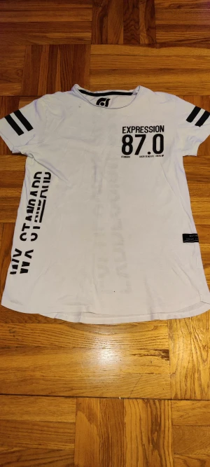 Vit t-shirt Expression 87.0 - Vit t-shirt med svart tryck 'EXPRESSION 87.0' framtill och grafiska texter längs sidan och ryggen. Svarta dubbla ränder på ärmarna ger en sportig vibe. Skön passform och rund halsringning. Perfekt för dig som gillar streetstyle.