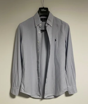 Ralph Lauren skjorta blå randig i S - Snygg blårandig skjorta från Ralph Lauren i custom fit. Skjortan har klassisk krage, knappar framtill och lång ärm med manschett. Ikoniska Polo-loggan broderad på bröstet. Tillverkad i mjuk bomull som känns skön mot huden.   Skjortan har inga defekter och är i ny skick🙌 hör av dig vid frågor/prisförslag😇🙌