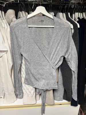 Grå omlottopp med knyt - Snygg grå omlottopp i storlek L. Toppen har långa ärmar och ett knytband i midjan som ger en cool och avslappnad look. Materialet är mjukt och stretchigt.