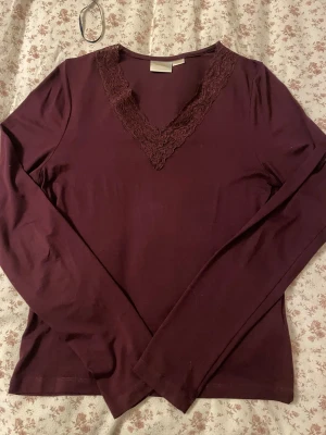 Vinröd långärmad topp från Gina Tricot - Säljer en vinröd långärmad topp från Gina Tricot med snygg spetsdetalj runt v-ringningen. Toppen har en slim passform och är tillverkad i mjuk bomull som känns skön mot huden. Perfekt för dig som gillar stilrena och feminina detaljer.