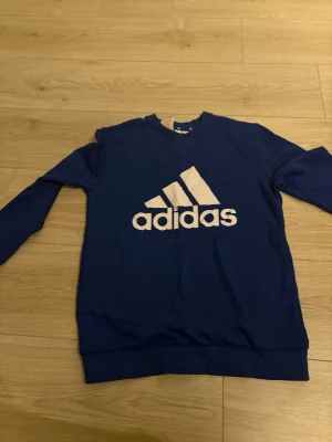 Blå Adidas sweatshirt med logga - Säljer en blå sweatshirt från Adidas med stor vit logga på bröstet. Tröjan har rund hals, långa ärmar och ribbade muddar vid ärmslut och nederkant. Materialet är mjuk bomull och passformen är normal. Perfekt för dig som gillar sportig stil.