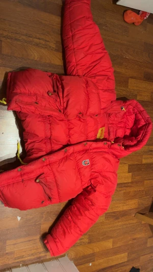 Röd dunjacka från Fjällräven S - Riktigt fet röd dunjacka från Fjällräven med huva och stora fickor. Jackan har stilren logga på bröstet, snörning i huvan och coola detaljer som tryckknappar och dragkedja. Perfekt för kalla dagar när du vill hålla stilen och värmen. Stor i storlek trots att den är i storken s. Köpt för 6000kr, pågrund av använt skick och fläckar vid fickan så säljer jag den lägre