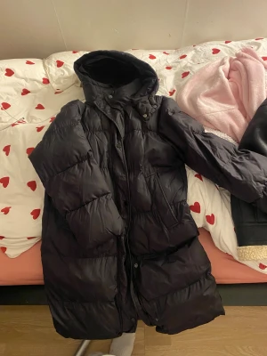 Svart lång pufferjacka med huva - Säljer en svart lång pufferjacka med huva och dragkedja. Jackan är vadderad och har en oversized look, perfekt för kalla dagar. Flera fickor framtill och hög krage som skyddar mot vinden. Snygg och praktisk vinterjacka för dig som vill hålla värmen.