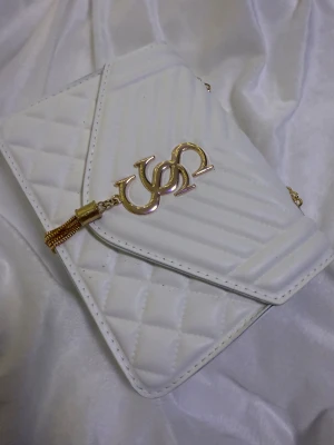 Vit quiltad axelväska med guldkedja - Snygg vit axelväska från U.S. Polo Assn. med quiltat mönster och guldfärgade detaljer. Väskan har en kedjerem i guld, magnetlås och dekorativt logohänge framtill. Tillverkad i syntetmaterial och har en stilren, rektangulär form.
