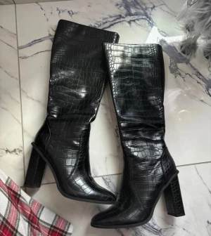 Svarta höga stövlar med krokodilmönster - Säljer ett par svarta stövlar med snyggt krokodilmönster och hög klack. Stövlarna har spetsig tå, dragkedja på insidan och är tillverkade i syntetmaterial med glansig finish. Perfekt för dig som vill sticka ut med ett coolt statement-plagg.