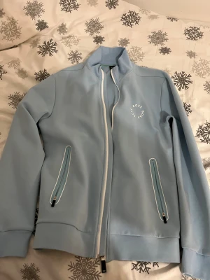 Ljusblå zipjacka från BOSS - Snygg ljusblå zipjacka från BOSS med vit dragkedja och logga på bröstet. Jackan har två fickor med dragkedja och ribbade muddar vid ärmslut och nederkant. Perfekt för dig som gillar stilren och sportig look.