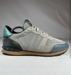 Valentino Garavani Rockrunner sneakers - Snygga Valentino Garavani Rockrunner sneakers i ljusgrå mesh och mocka med blå detaljer i skinn och gummisula. Ikoniska nitar på hälen och klassisk snörning framtill. Perfekt för dig som gillar exklusiva och stilrena sneakers med unik design.