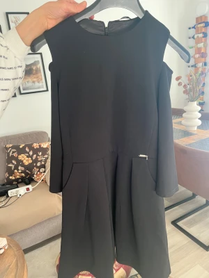 Svart kort klänning från Guess - Snygg svart klänning från Guess med markerad midja och lätt utställd kjol. Klänningen har långa ärmar med cold shoulder-detalj och dragkedja i ryggen. Stilren design med liten metallogo vid fickan. Perfekt för dig som gillar klassisk men trendig look.