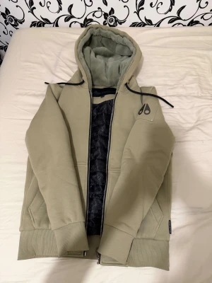 Beige hoodiejacka från Moose Knuckles - Snygg olivgrön/beige hoodiejacka från Moose Knuckles med svart logga på ärmen. Jackan har dragkedja, huva med mjuk fuskpäls på insidan och ribbade muddar. Insidan är quiltad och jackan har praktiska fickor framtill. Perfekt för kyliga dagar och fungerar som en hel vinterjacka. I princip nyskick