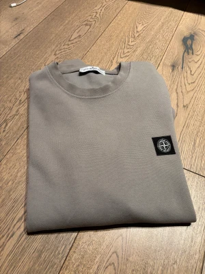 Beige sweatshirt från Stone Island - Snygg beige typ aktigt långarmad från Stone Island med klassisk rund hals och ikonisk patch på ärmen. Tröjan har en stilren design och är tillverkad i mjuk bomull som känns skön mot huden. Perfekt för dig som gillar streetwear och vill ha en clean look.