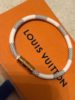  vitt armband med guldspänne - Snyggt armband  i vitt med svarta detaljer och ett exklusivt guldspänne där  är ingraverad. Armbandet har ett coolt mönster och känns lyxigt och trendigt, perfekt för dig som gillar designeraccessoarer.