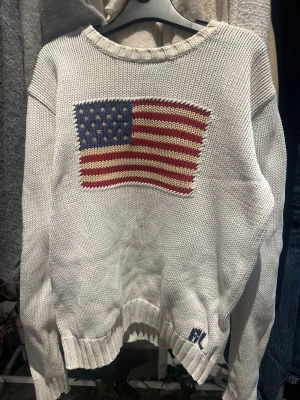 Vit stickad tröja Polo Ralph Lauren - Vit stickad tröja från Polo Ralph Lauren med amerikansk flagga broderad på bröstet och RL-broderi vid nederkanten. Tröjan har rund hals, ribbade muddar och lång ärm. Perfekt för dig som gillar klassisk preppy stil och vill sticka ut med en ikonisk detalj.