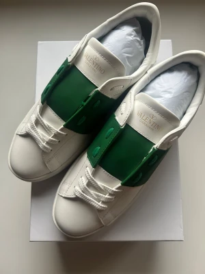 Valentino Garavani Open sneakers vit/grön - Äkta!! Snygga Valentino Garavani Open sneakers i vitt skinn med bred grön läderpanel över mitten. Klassisk rund tå, platt sula med nitar baktill och vita snören. Märkeslogga på plösen och stilren design som sticker ut med den gröna detaljen. Helt nya använt 1 gång!! Utan box 3499