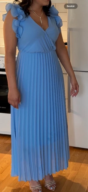 Ljusblå plisserad långklänning - Superfin ljusblå långklänning med plisserad kjol och volangdetaljer på axlarna. Klänningen har v-ringning och omlottliv, vilket ger en elegant känsla. Använde klänningen bara en gång. Fick många komplimanger för den 😍
