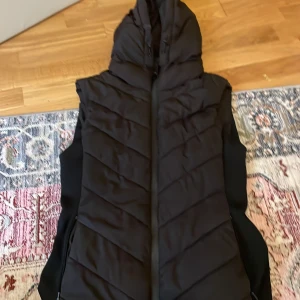 Svart quiltad dunväst från Zara - Svart dunväst från Zara med huva och dragkedja framtill. Västen är quiltad i ett snyggt mönster och har en stilren look. Perfekt att slänga över en hoodie eller tröja när det är kyligt ute.