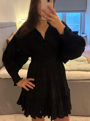 Svart kortklänning - Supersöt svart kortklänning med små vita prickar, v-ringning och knappar framtill. Klänningen har långa puffärmar och volangkant nertill. 