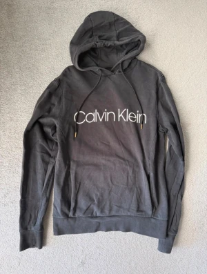 Svart Calvin Klein hoodie stl S - Snygg svart hoodie från Calvin Klein med stor vit logga på bröstet. Tröjan har huva med snörning och långa ärmar. Perfekt för chill dagar och enkel att matcha med jeans eller joggers. Jätte fint skick lite blekare än ny kanske. Rökfritt och djurfritt hem. 