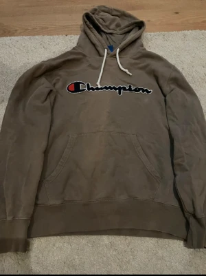 Brun hoodie från Champion strl L - Säljer en brun hoodie från Champion i storlek Large. Tröjan har klassisk huva med snörning, stor magficka och Champion-logga broderad på bröstet. Tillverkad i 100% bomull för skön och avslappnad känsla. Perfekt för chill dagar.