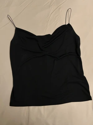  Ny Svart linnetopp från Gina Tricot - Svart linnetopp från Gina Tricot i storlek S. Tunn och mjuk polyester med smala axelband och lätt draperad front. Enkel och stilren design som är lätt att matcha med olika outfits. Perfekt för dig som gillar minimalistisk stil.