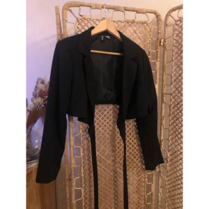 Svart croppad kavaj från DIVIDED - Snygg svart croppad kavaj från DIVIDED med långa ärmar och coola långa band framtill. Klassisk krage och stilren design som ger en modern vibe. Perfekt nu till alla festligheter i jul och nyår!