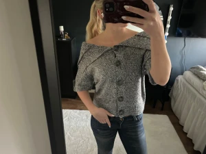 Grå offshoulder stickad topp med knappar - Säljer en gråmelerad stickad topp med offshoulder-design och stora knappar framtill. Toppen har korta ärmar och en bred krage som vilar snyggt över axlarna. Perfekt för dig som gillar en chill och trendig look.