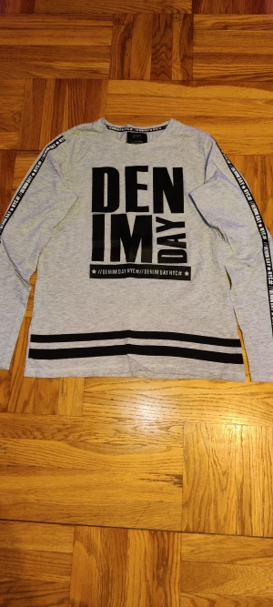 Grå långärmad tröja Denim Day NYC - Snygg grå långärmad tröja från Denim Day NYC med svart tryck framtill och svarta ränder längst ner. Svarta band med text längs ärmarna ger en cool streetkänsla. Rund hals och mjukt bomullsmaterial, perfekt för chill dagar. Skulle dock rekommendera att stryka den om man bryr sig mycket om sånt.