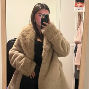 Beige pälsjacka med bälte - Säljer en beige pälsjacka med fluffig krage och muddar. Jackan har bälte i midjan för snygg passform och är oversized med långa ärmar. Perfekt för kalla dagar när du vill vara både varm och stylish.