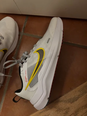 Nike sneakers vit/gul strl 38 - Nike sneakers i storlek 38 med vit mesh-ovandel, svart och gul swoosh på sidan och coola snörade band. Insidan är svart och yttersulan har detaljer i rött och blått. Perfekta för träning eller chill, med snygg sportig vibe och skön passform. Har aldrig använt dem. säljer billigt då jag inte vill ha de hemma.