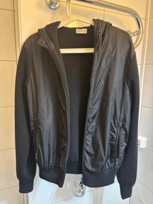 Moncler Cardigan   - Svart cardigan jacka från Moncler med huva och stickade ärmar. Framsidan har slät yta i syntetmaterial och två fickor med dragkedja. Moncler-logga på ärmen och ribbade muddar. Perfekt för dig som gillar stilren design och exklusiva märken. Moncler cardigan black label 