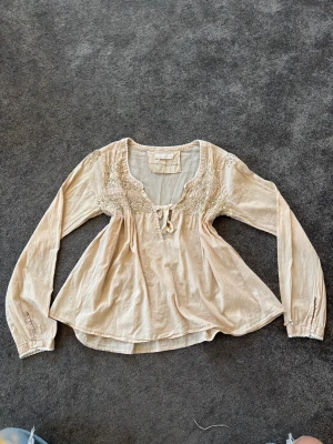 Odd Molly med paljetter - Supersöt beige blus från Odd Molly med broderade rosor och glittriga paljetter upptill. Blusen har långärmade ärmar, v-ringning med knytband och lätt volang i nederkant. 