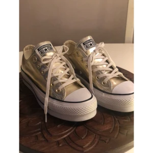 Guldiga Converse All Star, 38. - Snygga Converse All Star sneakers i metallic guld med vita snören och platåsula. Perfekta för dig som vill sticka ut med ett par glansiga och trendiga sneakers. Storlek: 38. Endast testade inomhus.