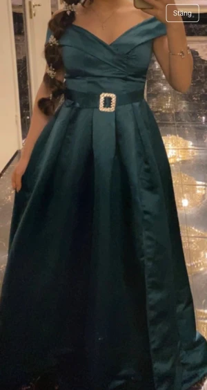 Mörkgrön långklänning med bälte - Snygg mörkgrön långklänning med offshoulder och v-ringad överdel. Klänningen har ett glansigt tyg och ett brett midjebälte med dekorativt spänne i pärlor. Kjolen är vid och faller elegant ner mot golvet, har en slits och passar perfekt för bal eller fest.