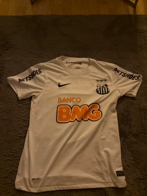 Santos Neymar Jr matchtröja Nike M - Säljer en äkta Santos FC fotbollströja från Nike, Neymar Jr #11. Tröjan är vit med svarta tryck, orange BMG-logga och klubbmärke på bröstet. Kortärmad, Dri-Fit-material för skön känsla och ventilation. Tryck på ärmarna och autenticitetsmärke. Perfekt för fans av brasiliansk fotboll.