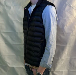 Marinblå Väst Tommy Hilfiger S - Svart pufferväst från Tommy Hilfiger i storlek S. Västen har quiltad design, ståkrage och dragkedja framtill. Materialet är syntet och polyester, med diskret logga på bröstet och klassiska Hilfiger-detaljer vid kragen. Perfekt att slänga över en hoodie eller tröja.