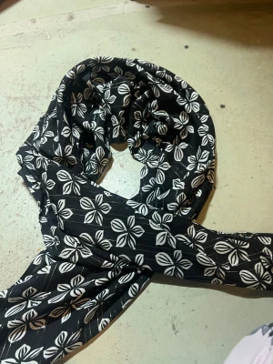 Svart blommig halsduk med vita detaljer - Snygg svart halsduk med vita blommor och tunna silverfärgade ränder. Tunt och mjukt tyg som känns lätt och luftigt, perfekt att slänga över axlarna för en cool look. Blommönstret ger en fräsch vibe och passar till många outfits.