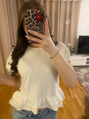 Vit blus med volang från Kappahl - Supersöt vit blus från Kappahl i mjuk bomull. Modellen har korta puffärmar och dubbla volanger längst ner som ger en gullig touch. Rund halsringning och liten smiley-detalj vid nederkanten. Perfekt för dig som gillar en clean och feminin stil.