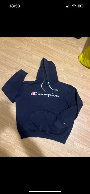 Marinblå Champion hoodie L - Säljer en marinblå hoodie från Champion i storlek Large. Tröjan har vit snörning vid huvan, stor logga på bröstet och klassisk känguruficka framtill. Mjuk bomullsmix och ribbade muddar vid ärmslut och nederkant. Perfekt för chill dagar.