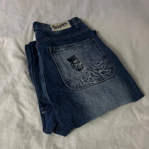 Derschutze blå jeans med broderi - Unika blå jeans från Derschutze med bred passform och coolt broderi av en tecknad figur på bakfickan. Jeansen har slitna detaljer och ljusare tvättade partier för en avslappnad vibe. Perfekt för dig som gillar streetwear och vill sticka ut.