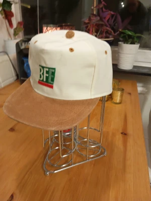 Vit och brun BFE keps med manchester - Snygg keps från BFE med vit krona och brun skärm i manchester. Framsidan har en broderad BFE-logga i grönt, vitt och rött. Kepsen har klassisk form och ventilation i toppen. Perfekt för dig som vill sticka ut med en unik stil.