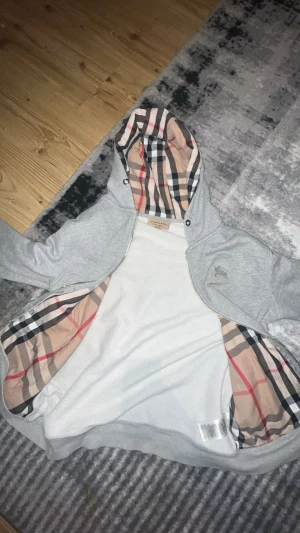 Grå Burberry hoodie med klassiskt mönster - Snygg grå hoodie från Burberry i storlek L men  dragkedjan är trasig går att fixa  och ikoniskt rutigt foder i huvan och insidan. Märkets broderade logga på bröstet och mjukt bomullsmaterial. Perfekt för dig som gillar exklusiv streetstyle och vill sticka ut med klassiska detaljer.